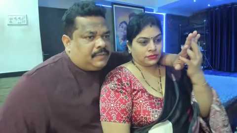 Media: Video of an Indian couple in a dimly lit room. The man, with a mustache, wears a maroon shirt; the woman, with short hair, dons a red sari with intricate designs.