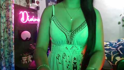 Media: Video of a woman with long black hair in a light green, patterned, lace-trimmed dress, standing in a dimly lit room with a neon \"Diva\" sign, colorful decor, and a bed in the background.