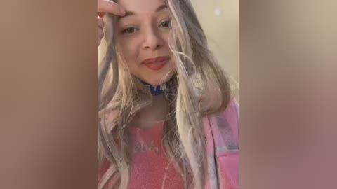 Media: Video of a young woman with long, wavy blonde hair, fair skin, and red lipstick, wearing a pink top and a blue choker, posing indoors.