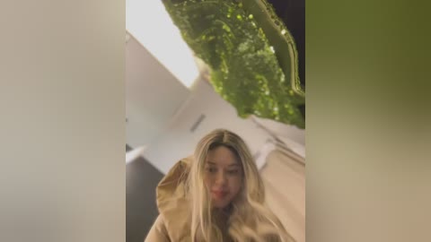 Video of a blonde woman in a beige coat, slightly blurred, in a modern room with a white wall and a large green plant in the background.