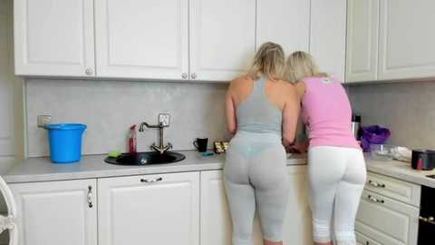 Media: Video of two women with blonde hair in tight, light-colored workout leggings and tank tops, washing dishes in a modern, white kitchen with gray countertops.