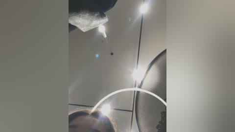 Media: Video of a person's head and neck, viewed from below, with a bright, lens flare effect; the background shows a ceiling with a white mesh fan and light fixtures.