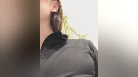 Media: Video of a woman with fair skin and medium-length brown hair, wearing a black collared shirt, captured from the neck up, against a blurred, bright background.