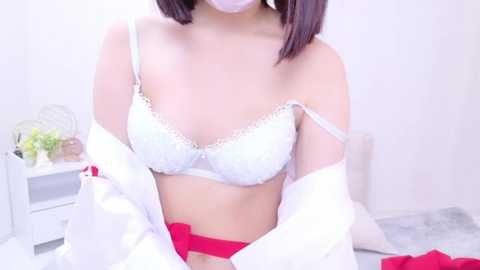 Media: A video of a light-skinned woman with straight, dark brown hair, wearing a white lace bra and red panties, partially unbuttoned white shirt, sitting on a bed in a bright, minimalistic room.