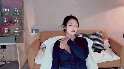 Media: A video of an Asian woman with fair skin, black hair in a bun, wearing a dark blue cardigan, lying on a bed in a minimalistic room with a TV displaying weather information.