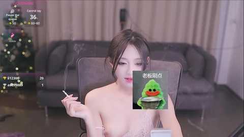 Media: Video of an East Asian woman with dark hair, topless, smoking a cigarette, sitting in a modern living room with a grey sofa and Christmas tree in the background.