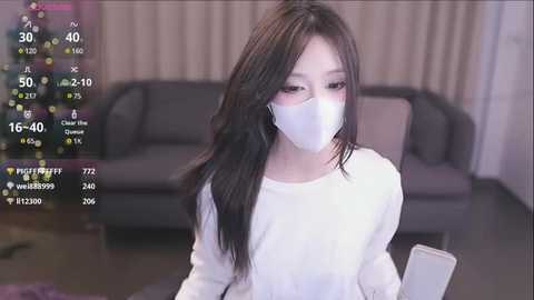 Media: Video of an East Asian woman with long black hair, wearing a white face mask and white sweater, holding a smartphone, indoors beside a gray couch.