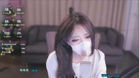 Media: Video of an East Asian woman with long black hair, wearing a white mask, white shirt, and glasses, sitting indoors near a grey sofa, with a digital temperature display.