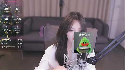 Media: Video of an East Asian woman with long black hair, wearing a white shirt, recording herself on a mic stand in a modern living room with a gray couch and Christmas decorations.