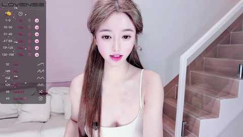 A video of a young Asian woman with long, straight brown hair, fair skin, and a slender physique, wearing a white tank top, standing in a modern, light-colored room with a staircase and white couch.