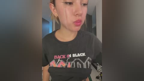 Video of a young Asian woman with blonde hair, wearing a black \"Back in Black\" AC/DC t-shirt, with mascara smudges and tears on her face, indoors.