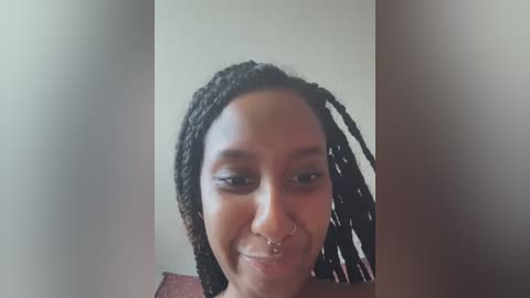 Media: Video of a smiling woman with medium-brown skin, wearing a nose ring and braided hair, against a plain beige background.