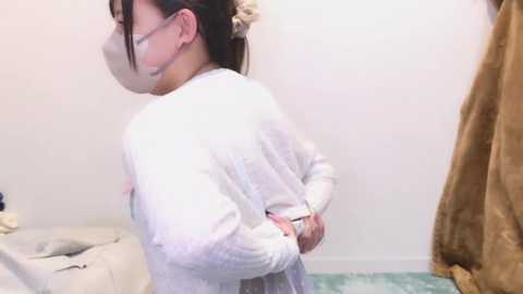 Media: Video of an Asian woman with a pale complexion, wearing a white lab coat, surgical mask, and glasses, standing in a hospital room with a white wall, a beige curtain, and a bed with white sheets.