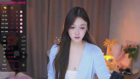 Media: A video of a young East Asian woman with long brown hair, wearing a light blue robe, in a dimly-lit room with a TV showing her face.