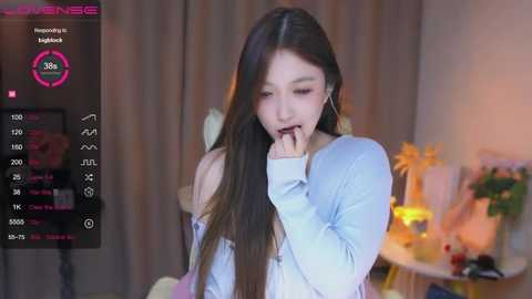 Media: Video of a young Asian woman with long black hair, wearing a light blue sweater, biting her finger in a casual indoor setting.