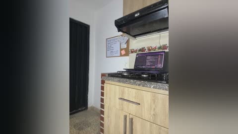 Media: Video of a compact kitchen with light wood cabinets, black stove, and a laptop on the counter, against a white wall with a corkboard and a small bulletin board.