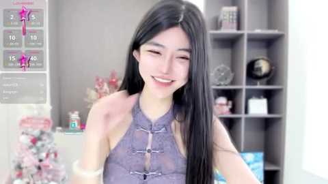 Media: Video of a smiling East Asian woman with long black hair, wearing a purple lace halter top, in a brightly lit room with bookshelves and decorative items.