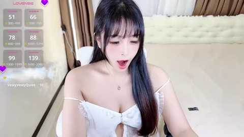 Media: A video of an East Asian woman with long black hair and pale skin, wearing a white off-shoulder top, sitting on a bed with a tufted headboard. The room has beige walls and a digital interface with heart icons and health stats.