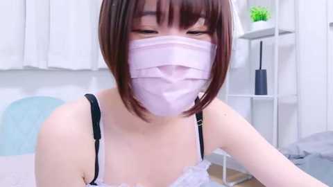 Media: Video of a young Asian woman with short brown hair, wearing a pink surgical mask, white lace bra, and black straps, sitting on a white bed in a minimalist room with a white wall and a small green plant.