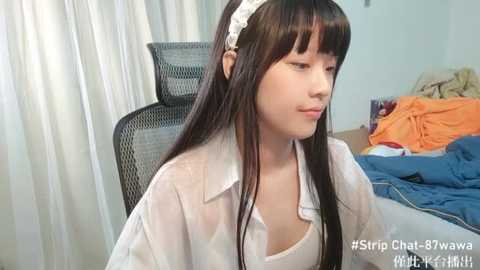 Media: Video of a young Asian woman with long black hair and a white headband, wearing a see-through white blouse, sitting in a chair amidst disheveled bedclothes in a messy bedroom.