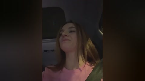 Media: A video of a young woman with straight, light brown hair, wearing a pink shirt, smiling slightly while seated in a car. The dim interior lighting casts shadows, with the car's dashboard visible in the background.