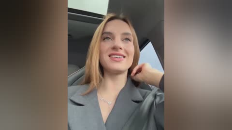 Media: Video of a Caucasian woman with straight blonde hair, smiling, wearing a gray blazer, inside a car with modern interior.