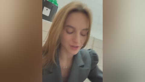 Media: A blurred video of a blonde woman in a gray blazer, partially obscured by a person's hand. The background shows a green plant and a white wall.