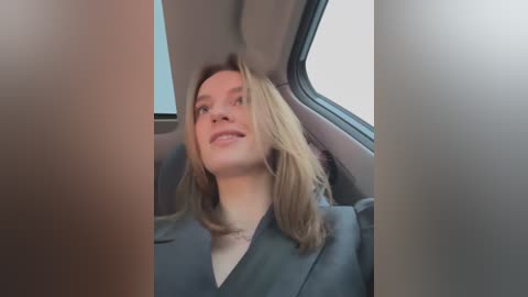 Media: Video of a smiling, fair-skinned woman with shoulder-length blonde hair, wearing a dark blazer, sitting in a car with gray interior.