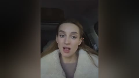 Media: Video of a Caucasian woman with straight brown hair, wearing a beige sweater, seated in a car with dim lighting, mouth open in surprise.