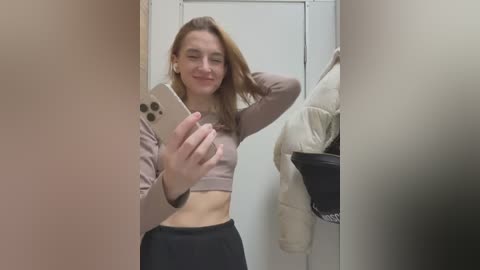 Media: A young woman with light skin and straight, light brown hair takes a selfie in a changing room, wearing a cropped, light purple top and black pants.