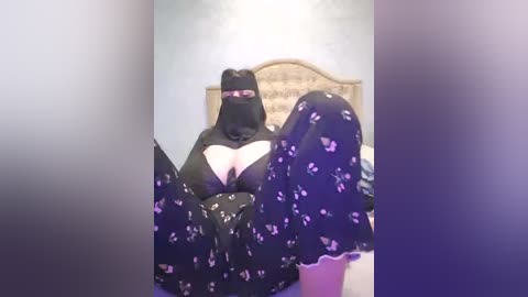 A video captures a woman wearing a black face mask and a black top with a cut-out design, revealing ample cleavage, and black pajamas adorned with purple butterflies. She is seated on a bed with a beige headboard in a softly lit room.