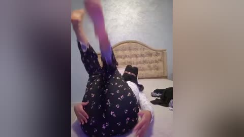 Media: A video of a person lying on their back on a bed, wearing black pajama pants with white floral patterns, legs raised and hands on their buttocks, in a bedroom with a beige upholstered headboard and light blue walls.