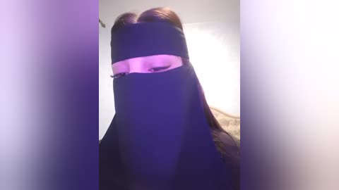 A video of a person wearing a black burqa with a pink strip over the eyes, standing in a dimly lit room with a white wall.