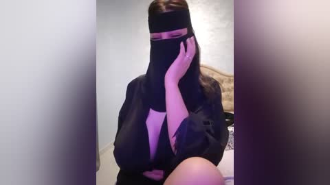 Media: Video of a person wearing a black niqab, with long dark hair, sitting in a room with beige walls and a bed visible in the background. The image is blurred, with a purple tint.