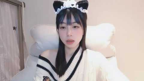 Media: Video of an Asian woman with long black hair wearing a black and white maid outfit, including a cat ear headband, seated in a white chair. Background includes a wooden door and beige wall.