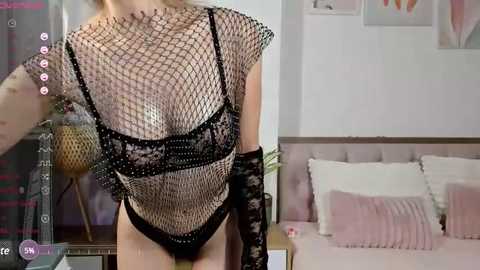 Media: Video of a woman wearing a black fishnet bodysuit and lace gloves, standing in a modern bedroom with pink accents and framed art on the wall.