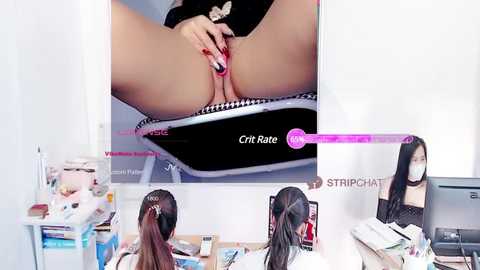 Media: A video showing two women with long hair, one using a pink vibrator on her vulva, seated at a desk with a computer monitor, surrounded by office clutter.