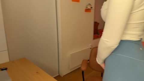 Media: Video of a woman in a cream sweater and blue pants, standing in a small, brightly lit room with beige walls, a wooden desk, and a chair.