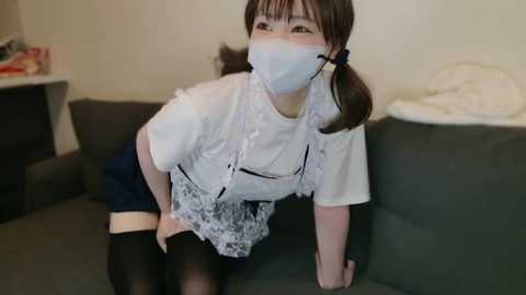 Media: A young Asian woman with fair skin, brown hair, and bangs, wearing a white lace dress, black stockings, and a white face mask, leans on a grey couch in a dimly lit room.