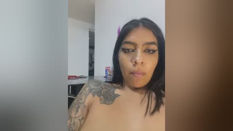 Media: A video of a young woman with medium skin tone, black hair, and a tattoo sleeve, puckering her lips in a selfie. The background shows a modern kitchen with white walls and cabinets.