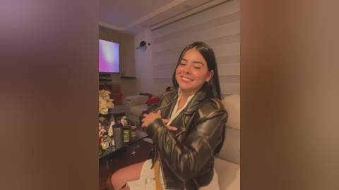 Media: Video of a young Latina woman with long black hair, wearing a black leather jacket, white top, and white shorts, smiling, in a cozy living room with a TV, floral arrangement, and a couch.