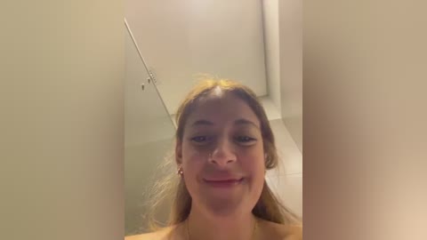 Media: Video of a young woman with light skin, light brown hair, and a slight smile, standing in a shower with a white tiled wall. She wears a yellow shirt.