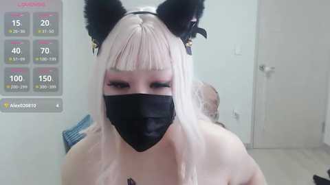 Video of a young Asian woman with light skin, platinum blonde hair, and black cat ear headband. She wears a black face mask, pink eyeshadow, and a black top. Background features a white door and a blue cushioned chair.