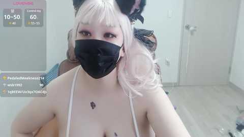 Media: Video of a fair-skinned woman with pastel pink hair and black cat ears, wearing a black face mask and revealing top, taken indoors with a live streaming overlay.