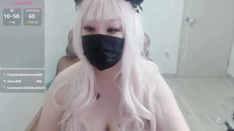 Media: A video of a pale-skinned woman with long, wavy platinum hair, wearing a black face mask, black headband, and a sheer pink top, in a minimalist room with white walls and a gray floor.