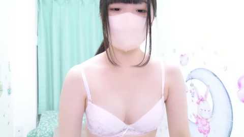 A video of an Asian woman with straight black hair, wearing a pink lace bra, a face mask, and a white shirt. She stands in a brightly lit room with green curtains and floral wallpaper.