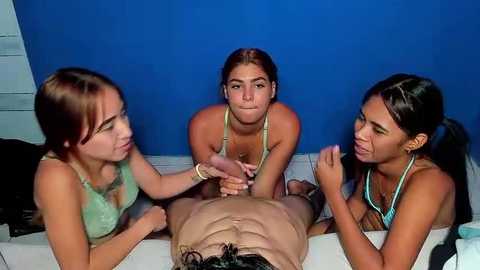 Media: Video of three teenage girls with light skin and brown hair, wearing green tank tops, kneeling around a prone man with dark hair, in a blue room.