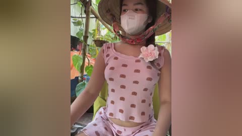 Media: Video of a young woman with light brown skin and long hair, wearing a pink dress with a heart pattern, a floral headband, and a face mask, seated among lush greenery.