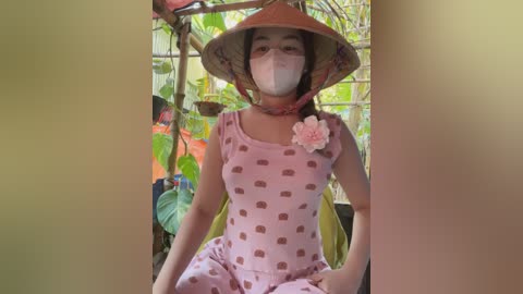 Media: Video of an Asian woman with light skin and medium-sized breasts, wearing a pink dress with heart patterns, a pink flower on her chest, and a conical hat, seated in a green, lush garden.