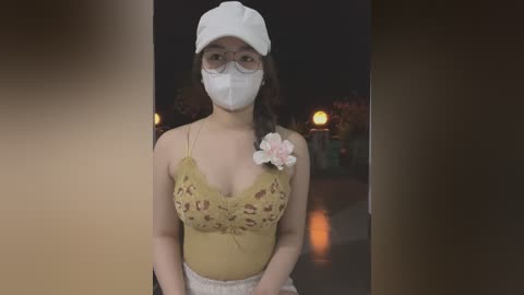 Media: Video of a young woman with light brown skin and dark brown hair, wearing a white mask, glasses, and a mustard-yellow lace top with a pink flower. She stands in a dimly lit, blurred background.